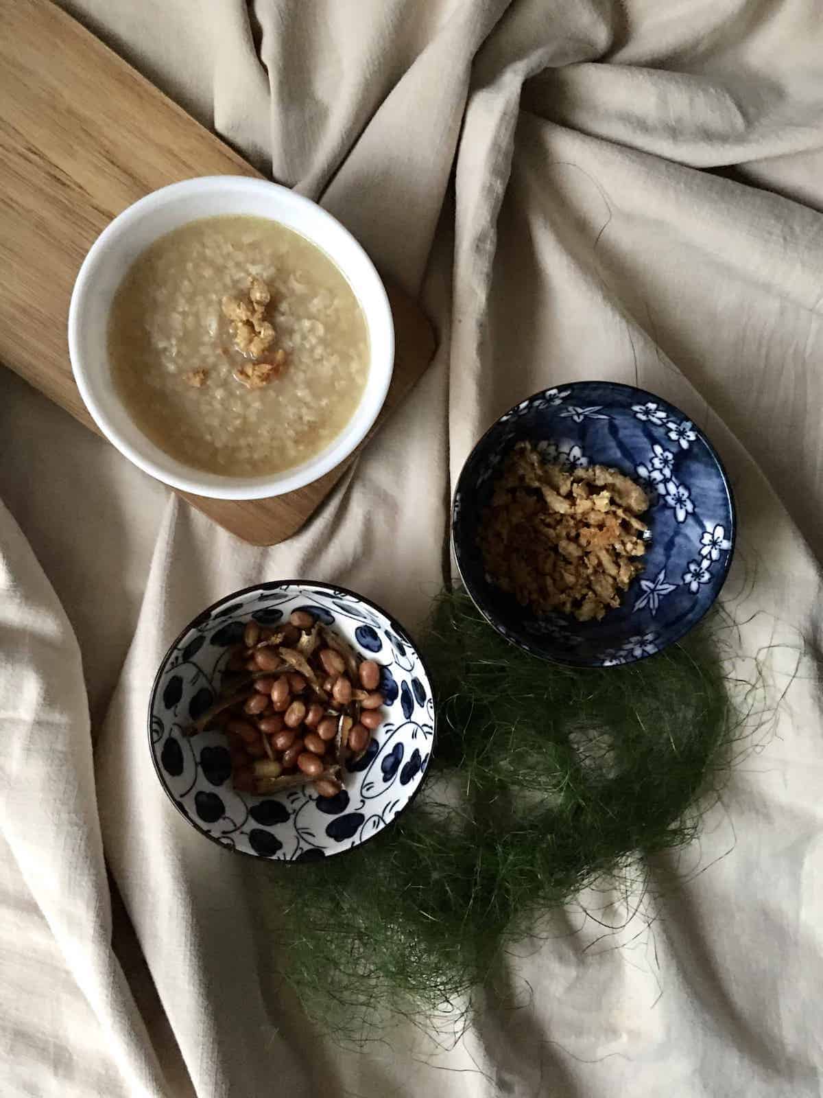 A bowl of Singaporean Hainanese Chicken Porridge topped with crispy chicken skin, with fried peanuts and pork crackling on the side