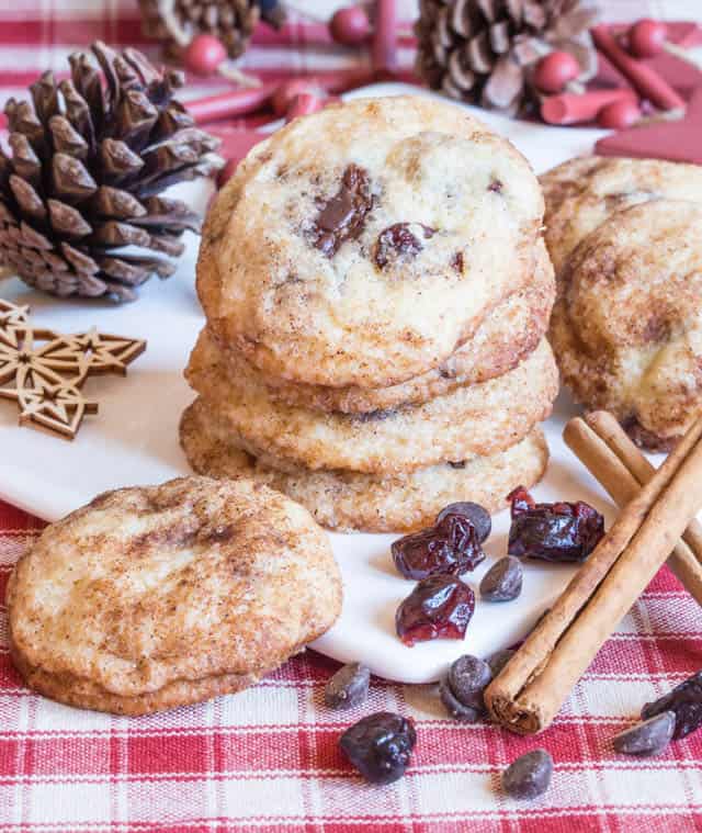A stack of cranberry and chocolate snickerdoodle cookies with cranberries, cinnamon and a pinecone piled around