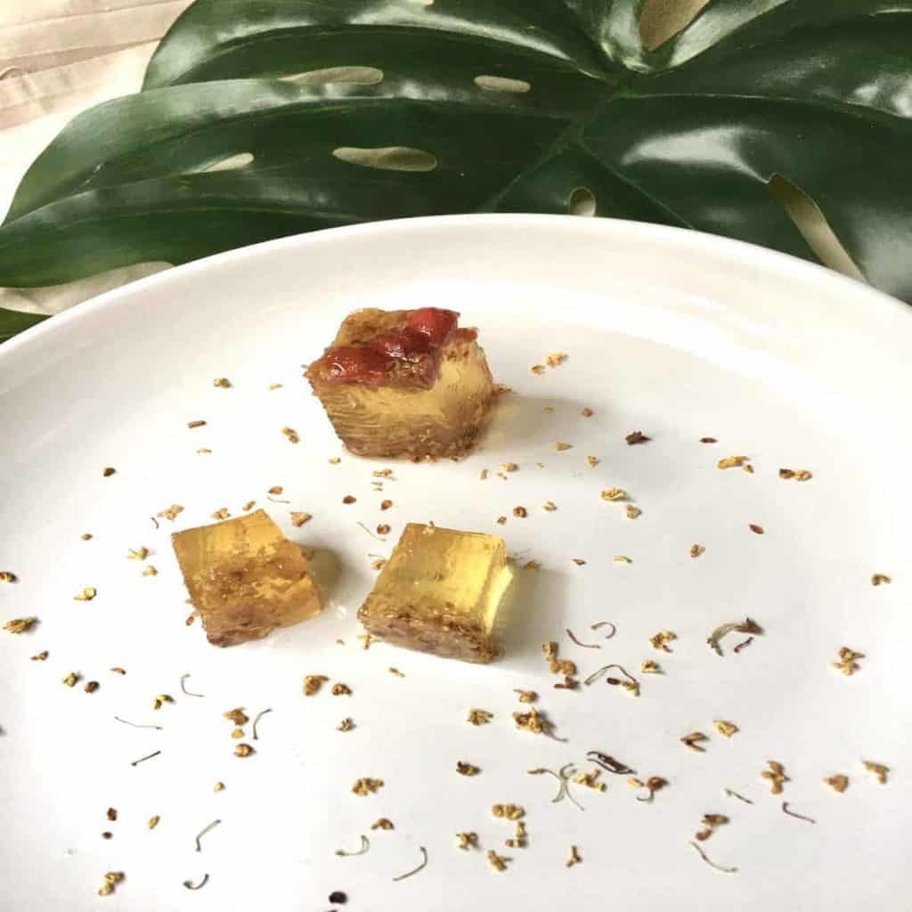 3 golden cubes of Osmanthus tea jelly on a plate with Osmanthus flower tea sprinkled around it to create a pretty plate