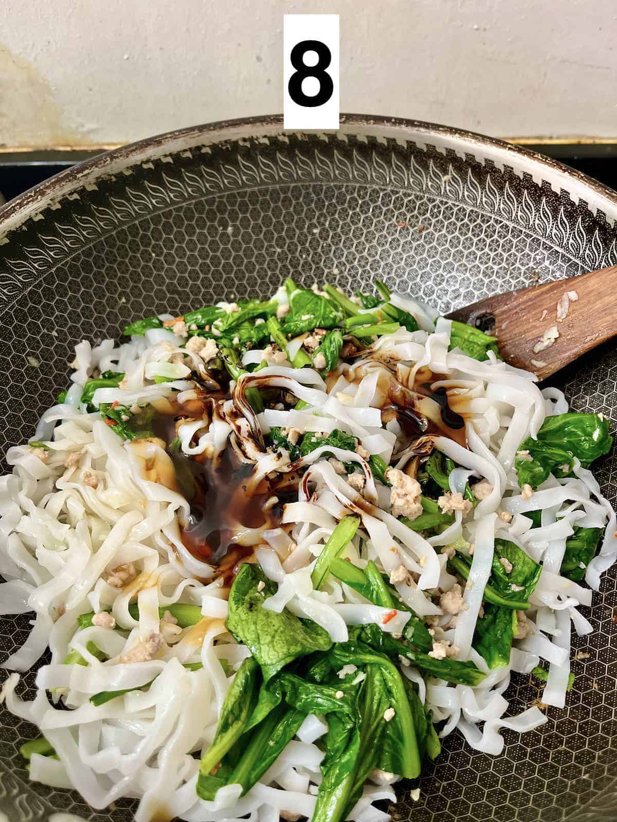 Rice noodles, green vegetables and pork with sauces on top, in a wok.