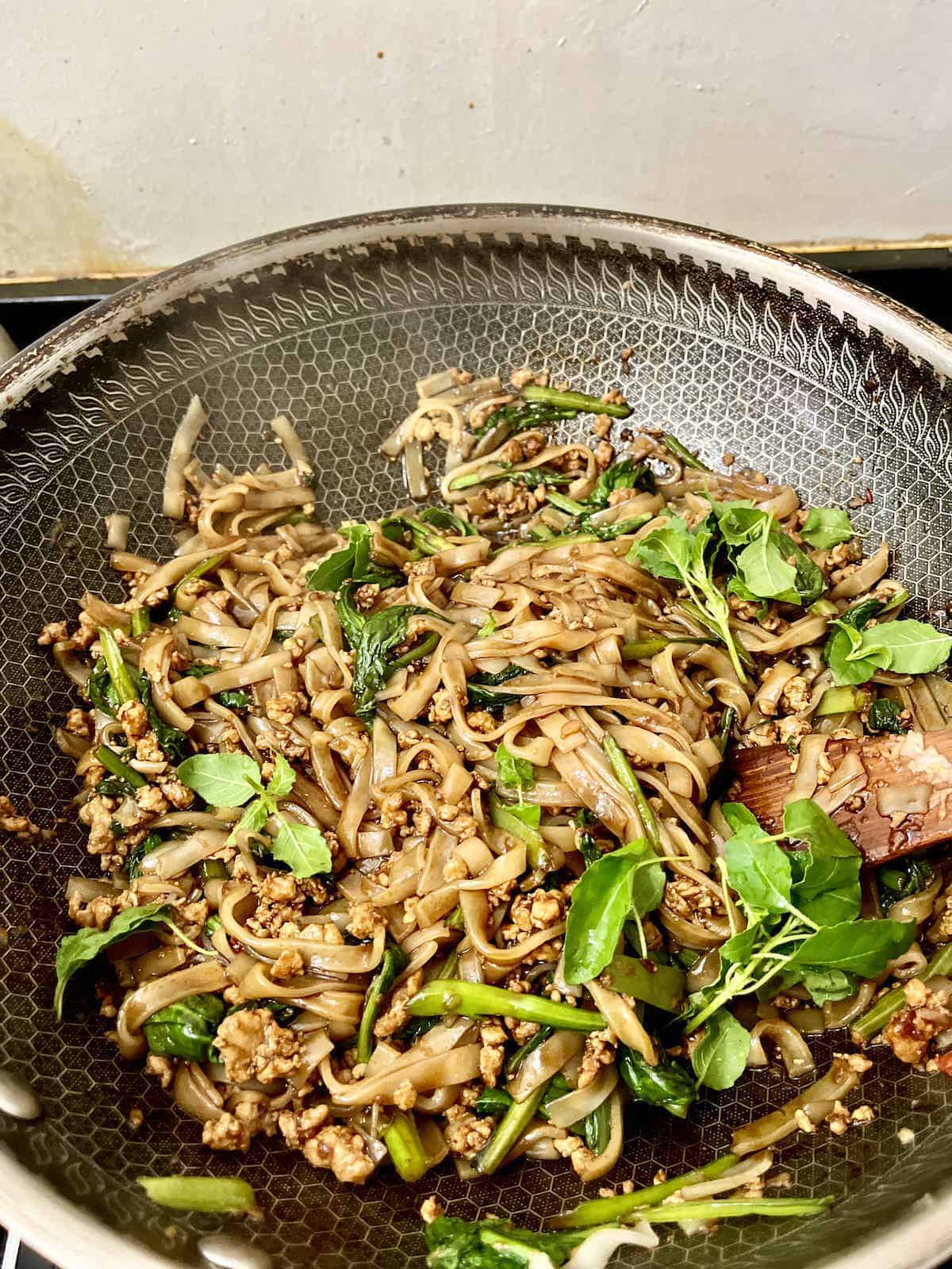 Adding holy basil leaves to a wok full of dark noodles.