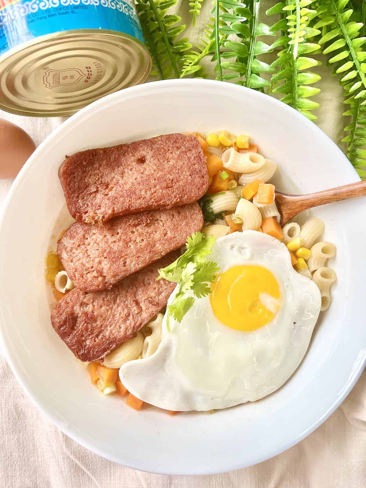 A bowl of macaroni with a fried egg, corn, carrots and luncheon meat.