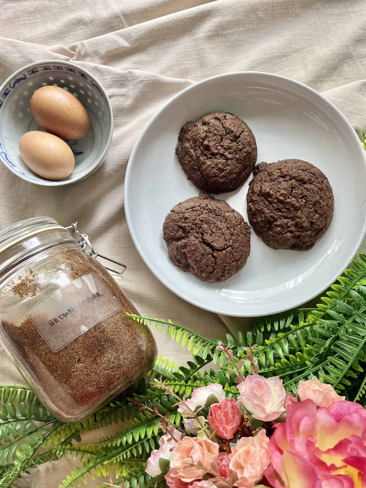 3 black sesame chocolate cookies next to eggs and brown sugar.