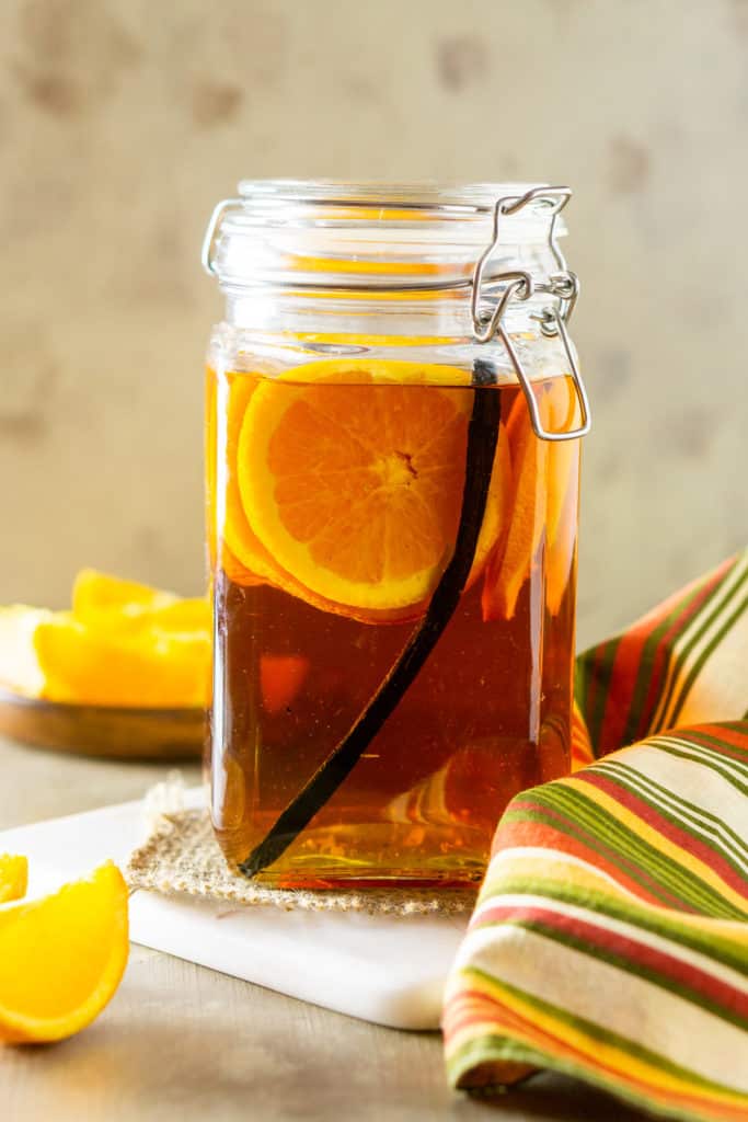 A large jam jar full of bourbon with some orange slices and a vanilla bean inside