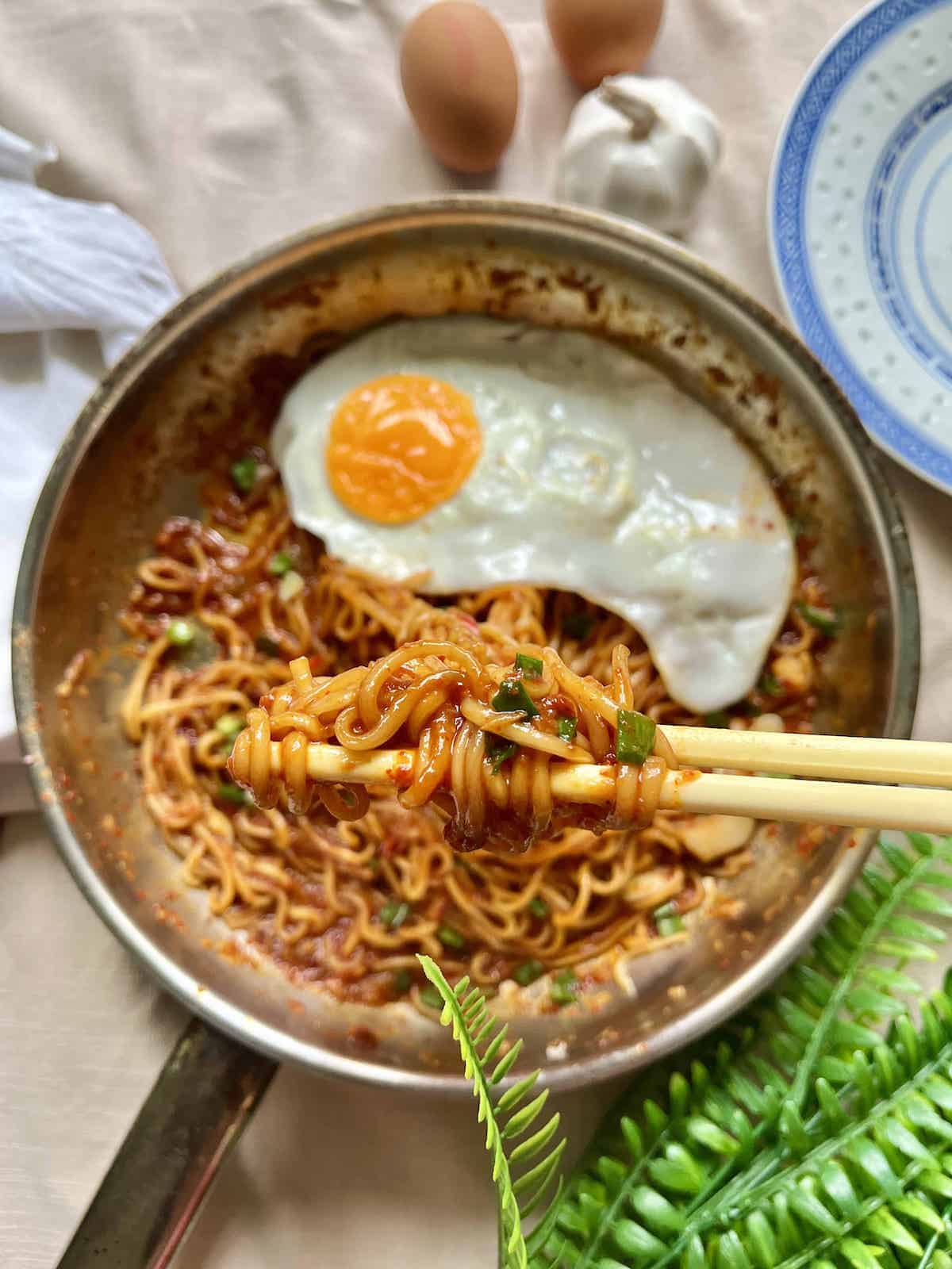 A pair of chopsticks lifting spicy noodles off a pan.