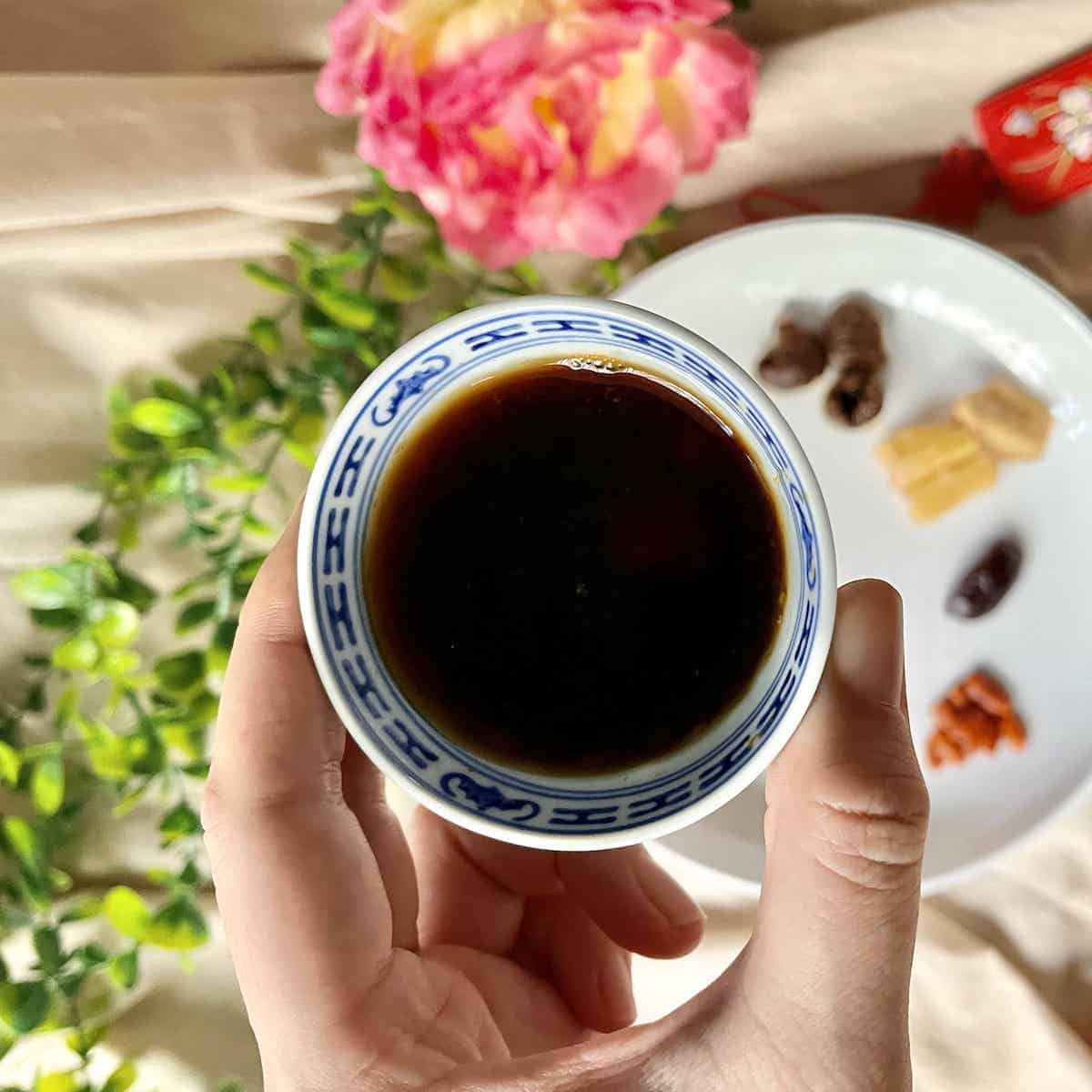 Someone holding a cup of longan jujube tea with other TCM ingredients in the background.