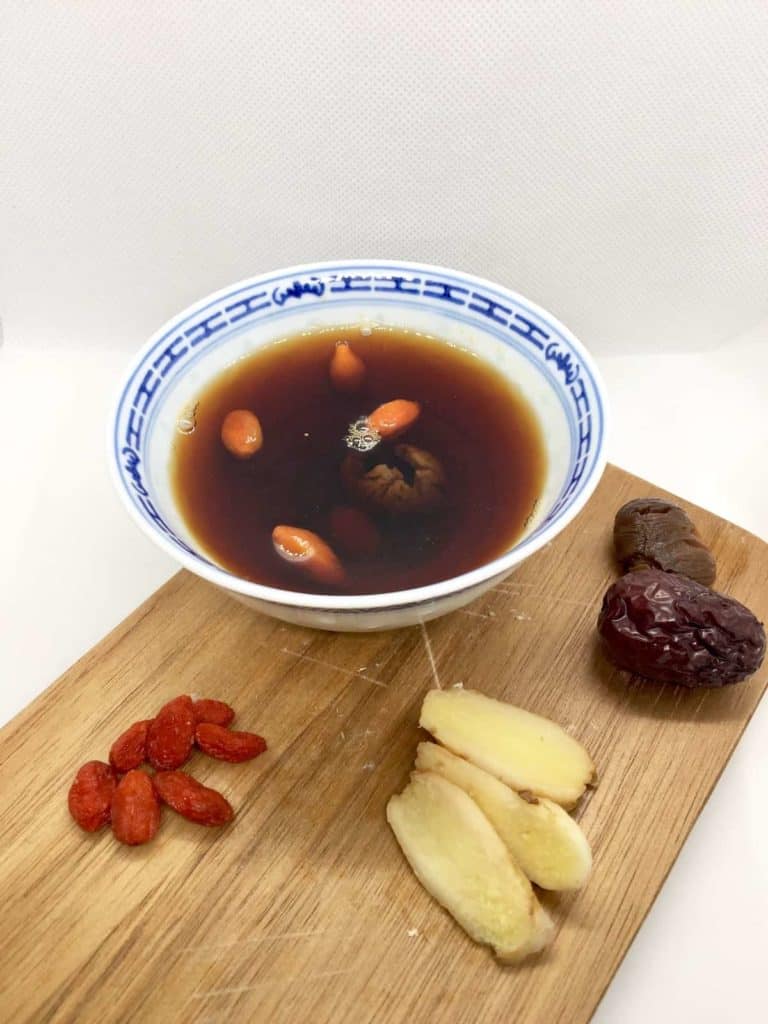 A cup of dried longan tea with wolfberries in it and goji berries, ginger slices and jujube around it