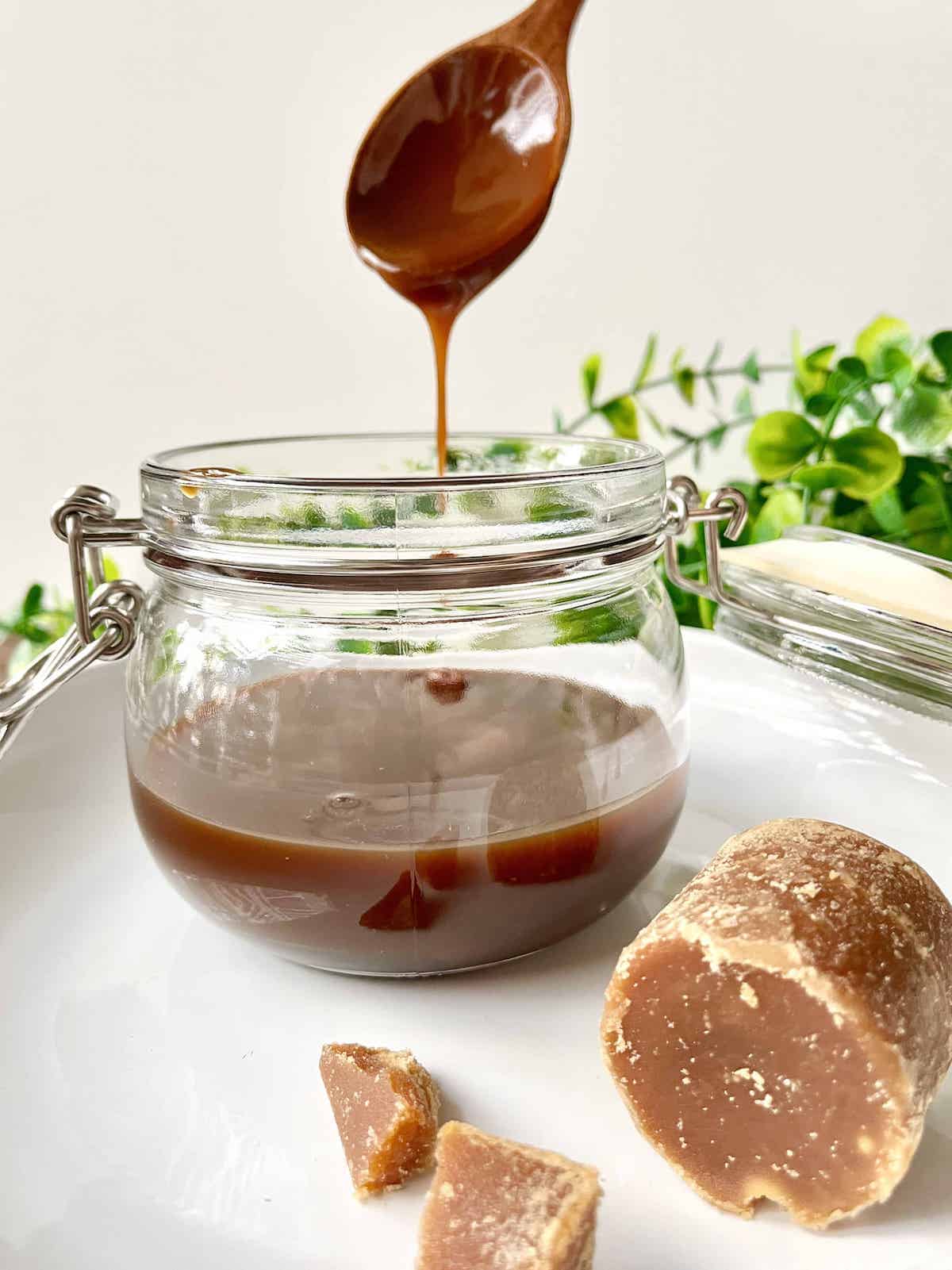 A wooden spoon scooping coconut caramel sauce out of a jar next to Gula Melaka Palm Sugar.