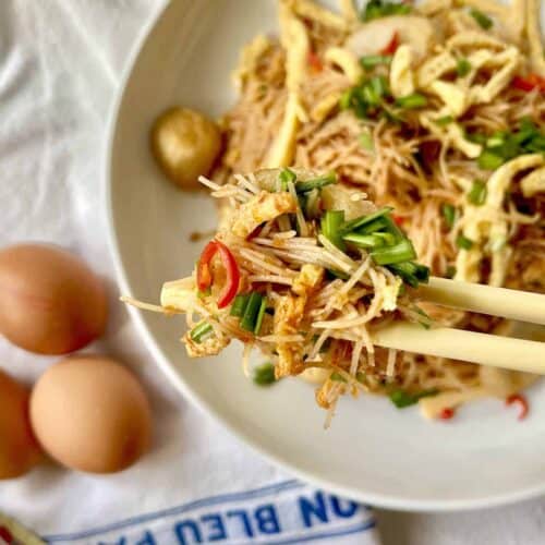 A pair of chopsticks grabbing some fried mee siam goreng with eggs and red chili.