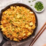 A skillet brimming over with noodles cooked in a peanut sauce and tofu