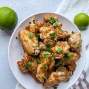 A pile of brown chicken wings with spring onions on a plate and 2 limes next to it