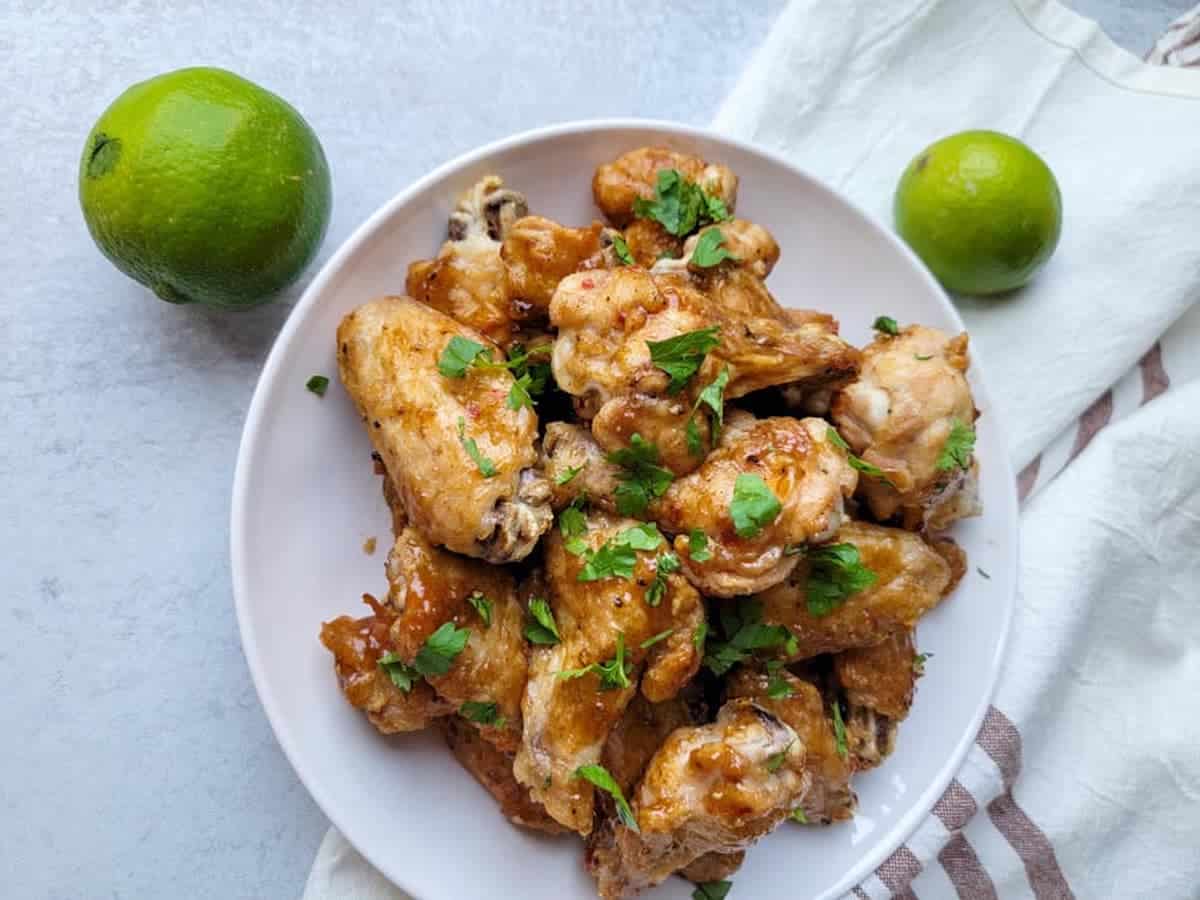 A pile of brown chicken wings with spring onions on a plate and 2 limes next to it