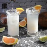 2 glasses of coconut daiquiri with fresh coconut, grapefruit and lime next to them.