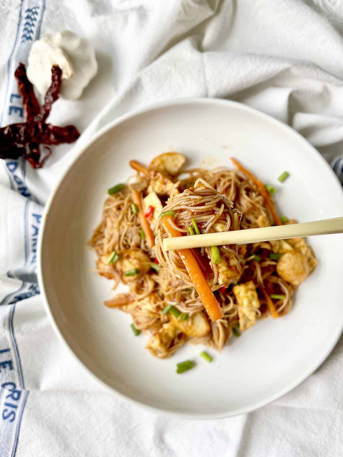 A pair of chopsticks grabbing some fried vermicelli noodles with sliced carrots.