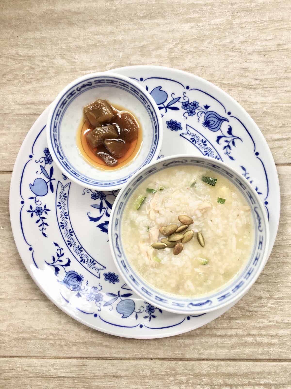 A bowl of rice porridge topped with toasted pumpkin seeds and Chinese pickled lettuce on the side.