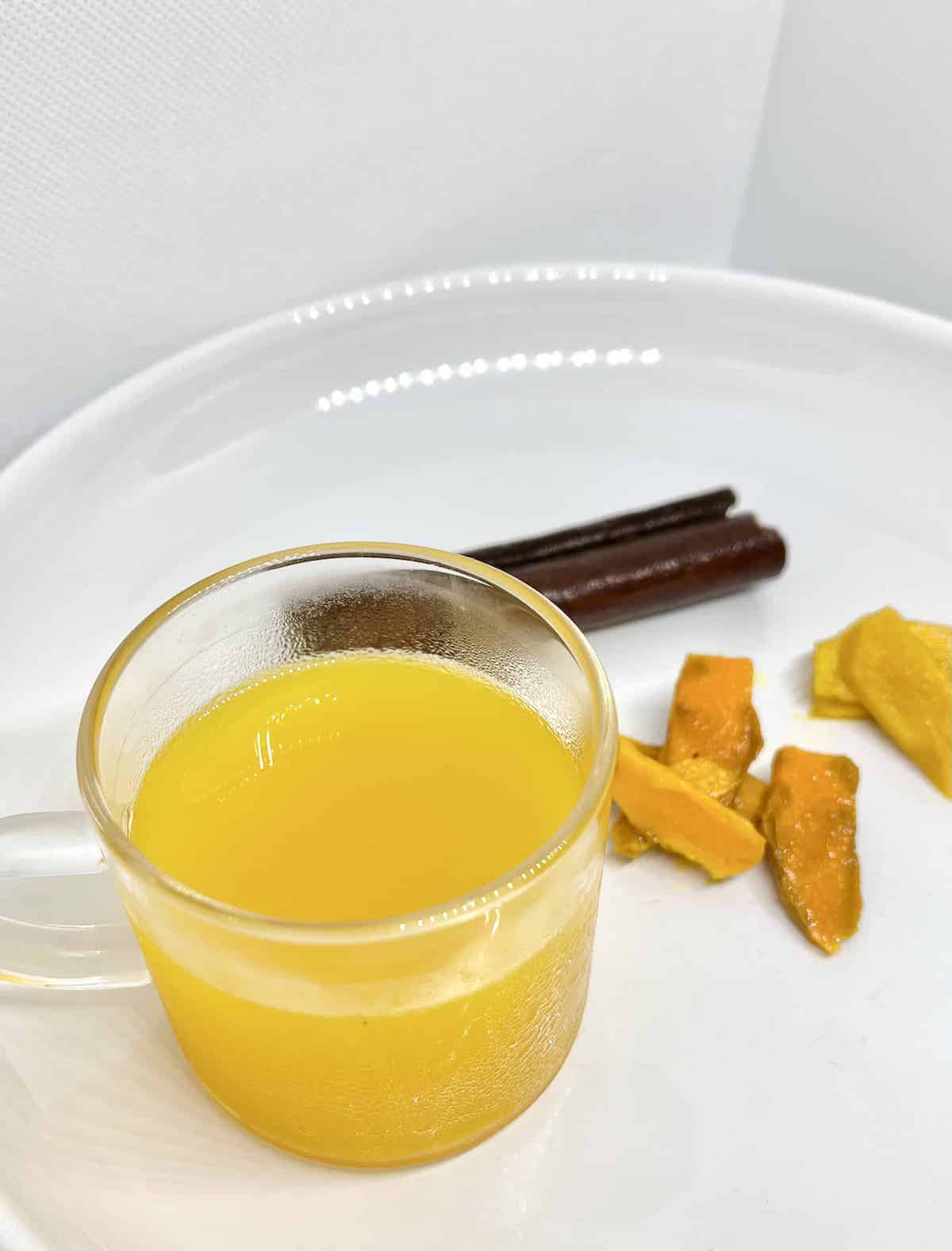 A glass of golden tea with turmeric, ginger and cinnamon behind it.