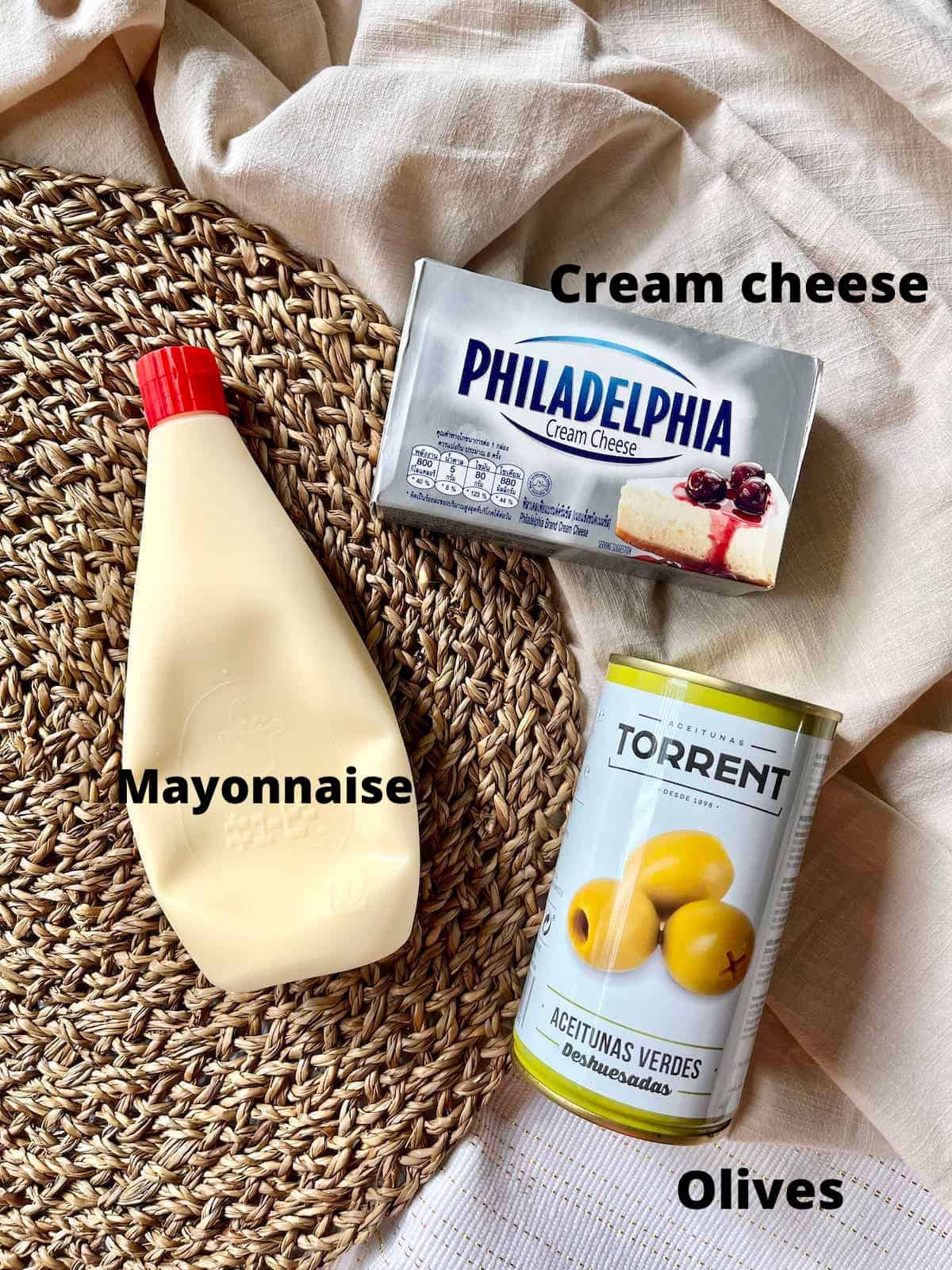 The 3 ingredients needed to make olive cream cheese spread on a tablecloth.