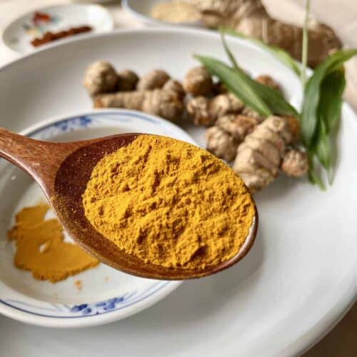 A close-up of a spoonful of dried turmeric powder with fresh turmeric in the background.