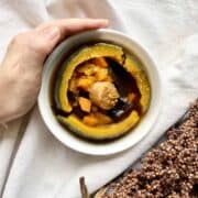 A hand holding Chinese pumpkin soup served in a small pumpkin and white bowl.