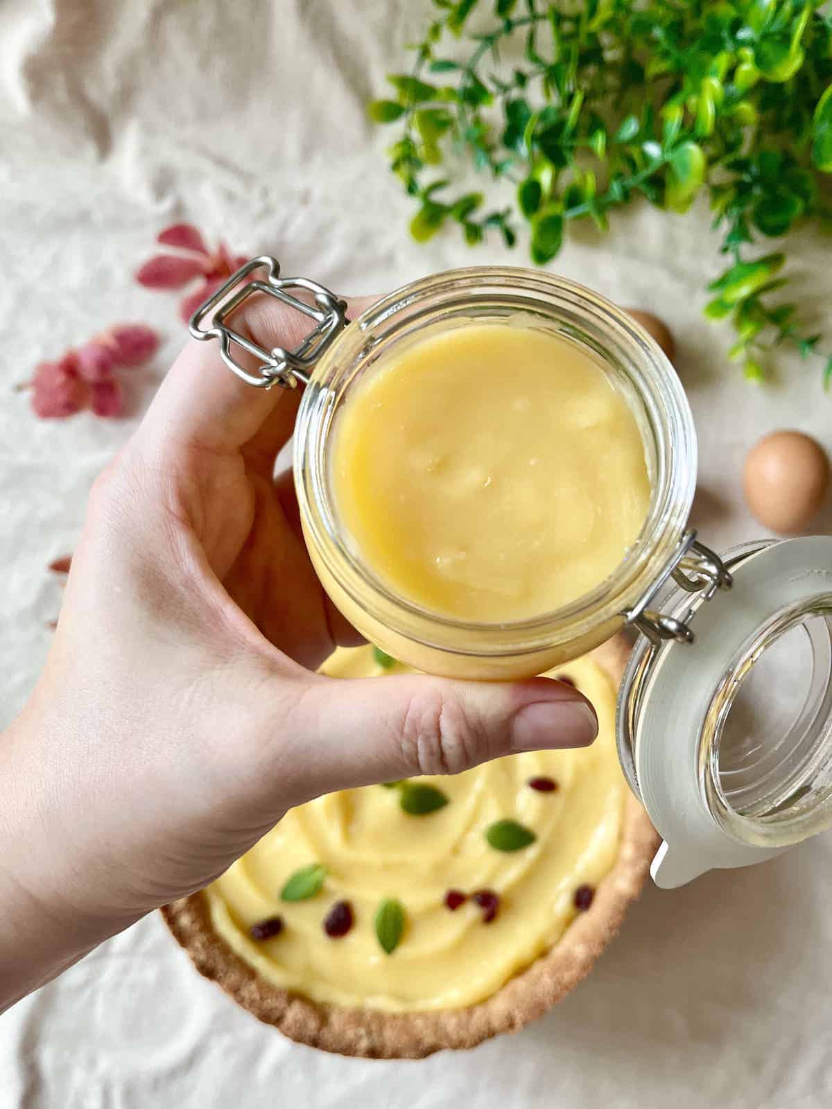 Someone holding a jar of lime curd with a lime curd tart in the background.