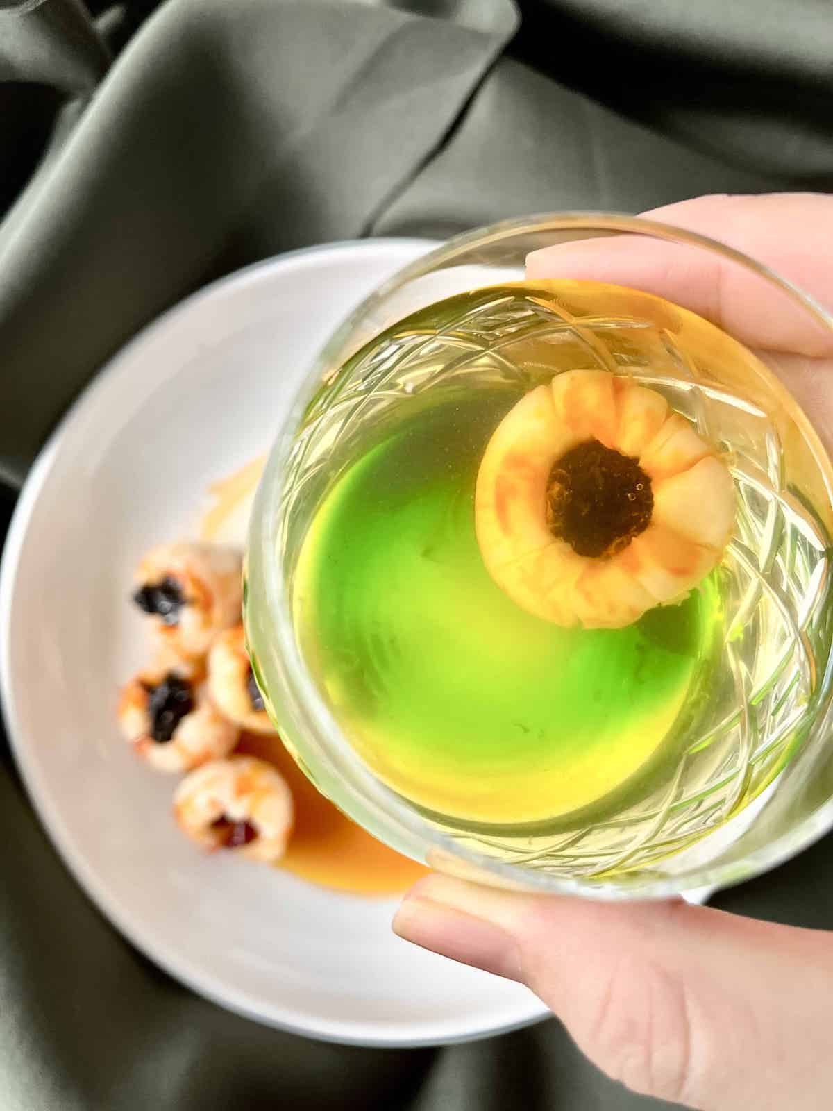 A cup of odiously green cocktail with a bloody looking lychee eyeball floating in it.