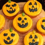 5 orange cupcakes decorated as Jack O Lanterns on a wooden log board.