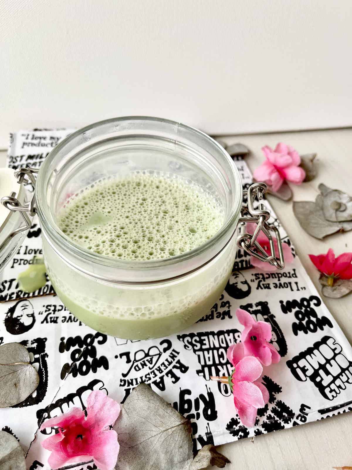 A mason jar green latte made with oat milk and matcha surrounded by pink flowers.