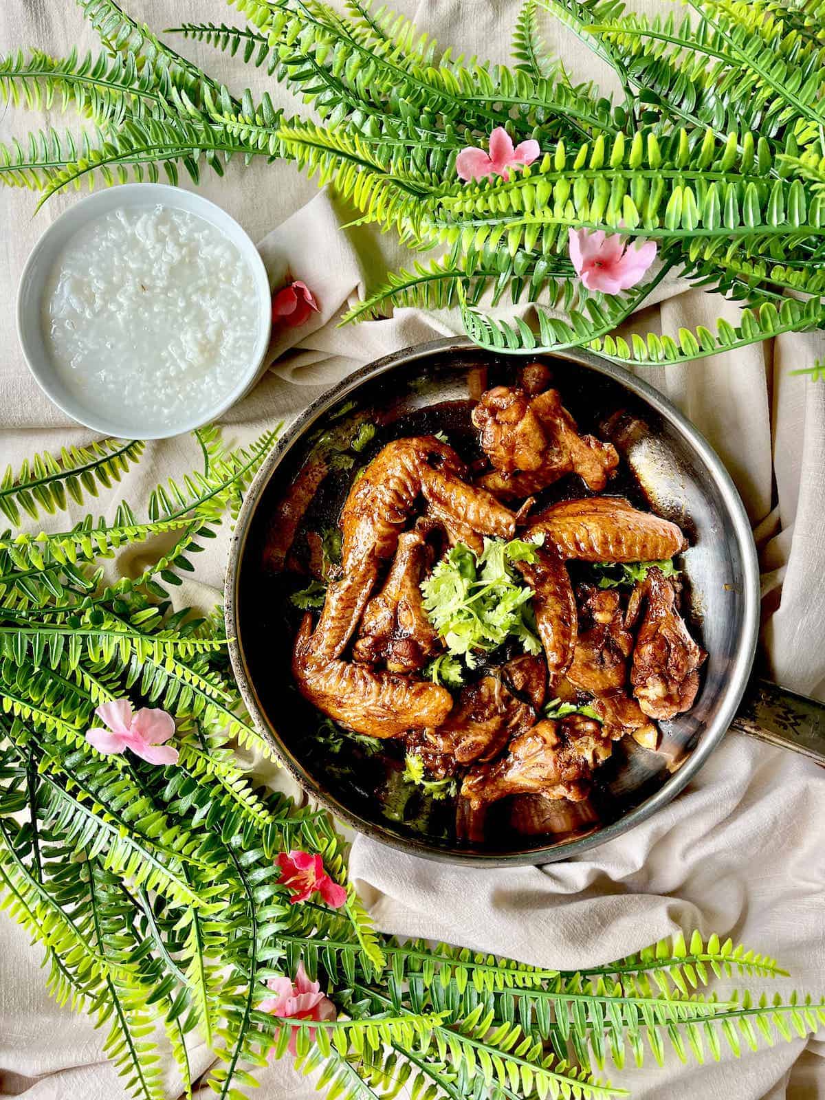 A plate full of sticky Chinese chicken wings next to a bowl of rice porridge.