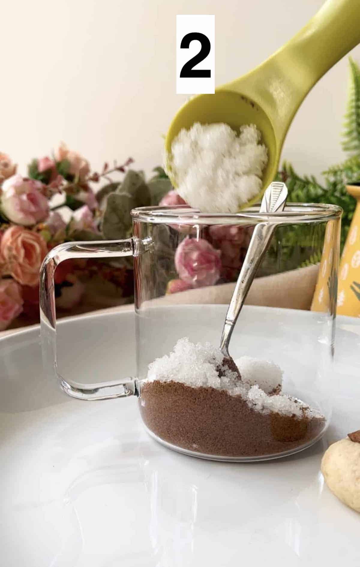 Adding white sugar to milo powder in a glass cup.