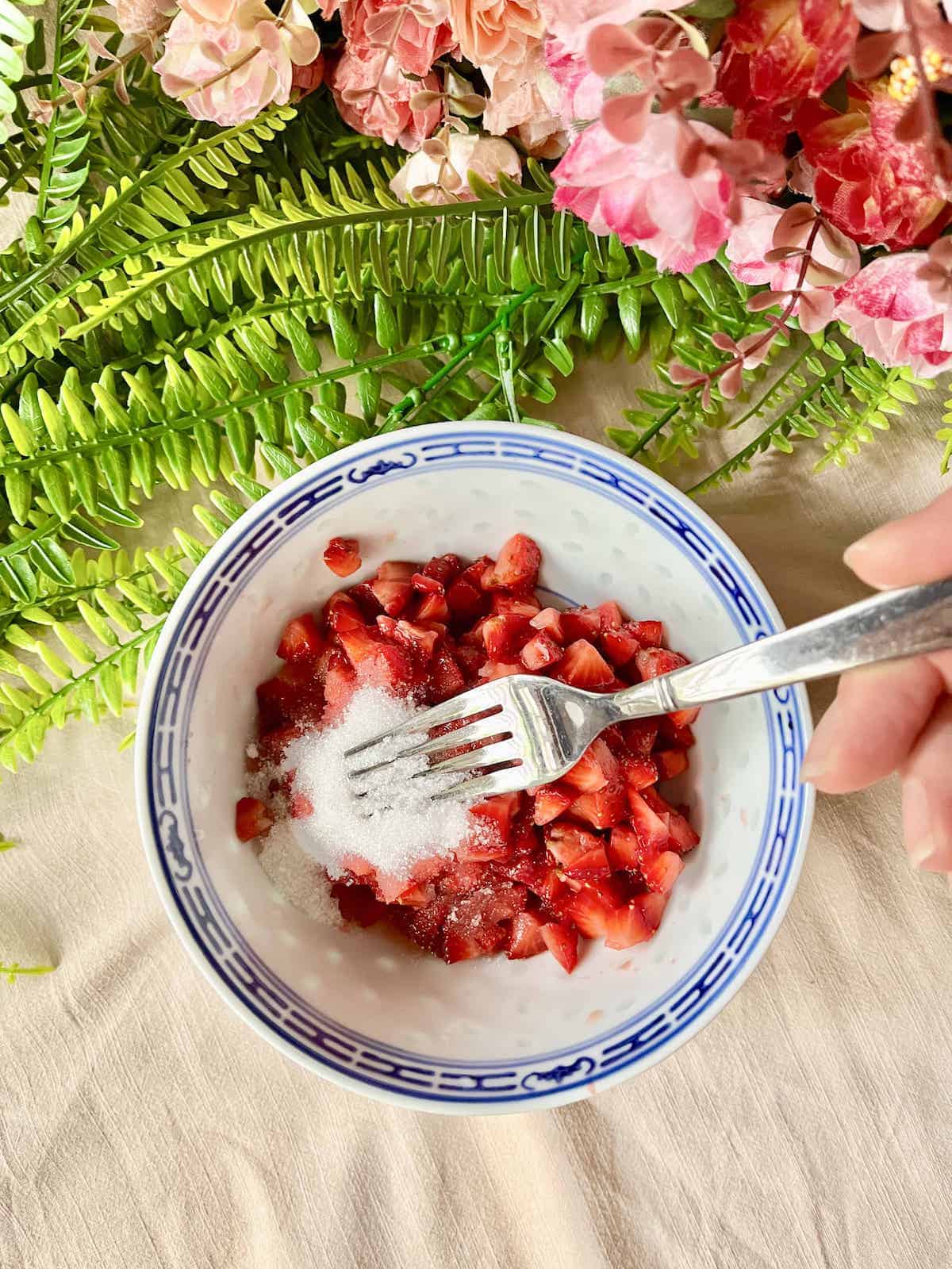 Mashing cut up strawberries with white sugar using a fork.
