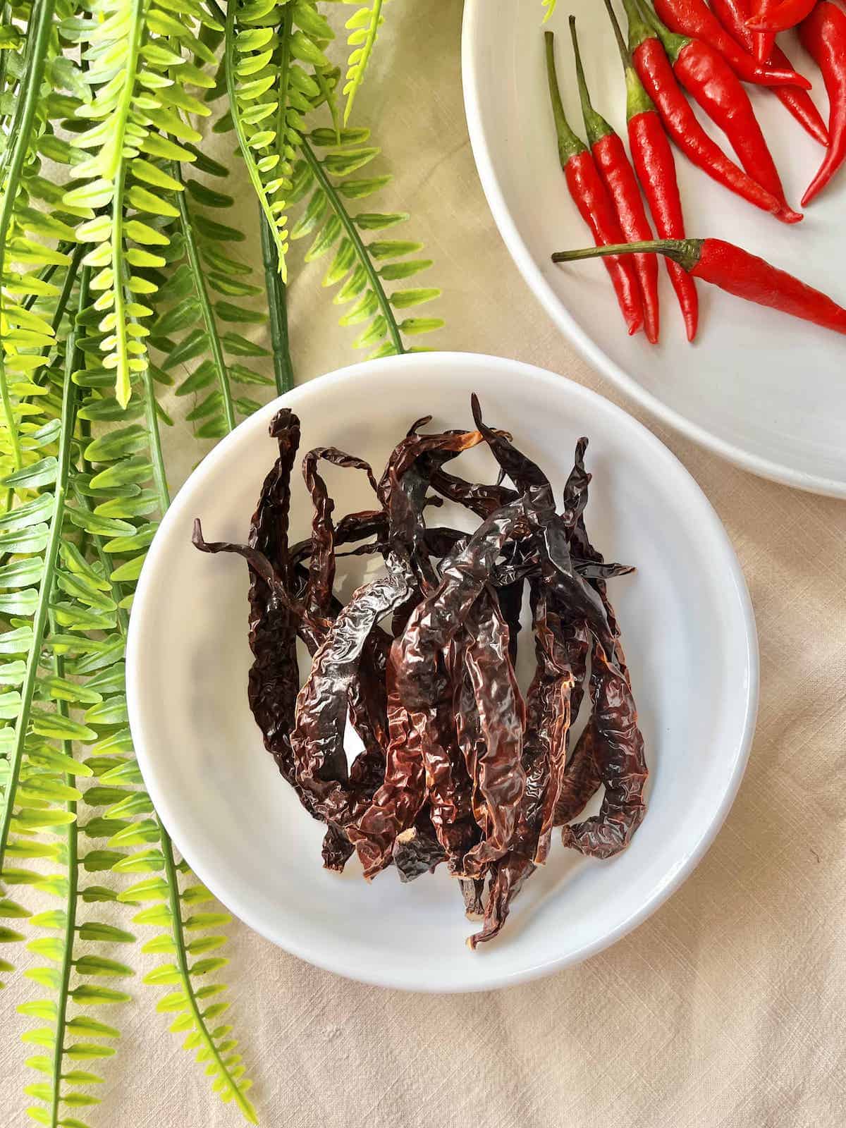 A bowl of dried chilies next to a plate fo fresh Thai Chilies.