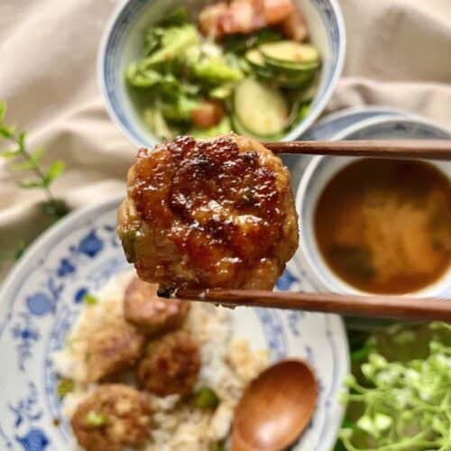 A wooden chopstick picking up a Japanese chicken teriyaki meatball from a plate of rice.