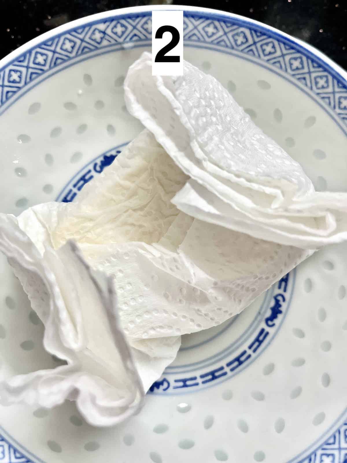 A block of tofu wrapped in a paper towel and placed on a blue and white Chinese plate.