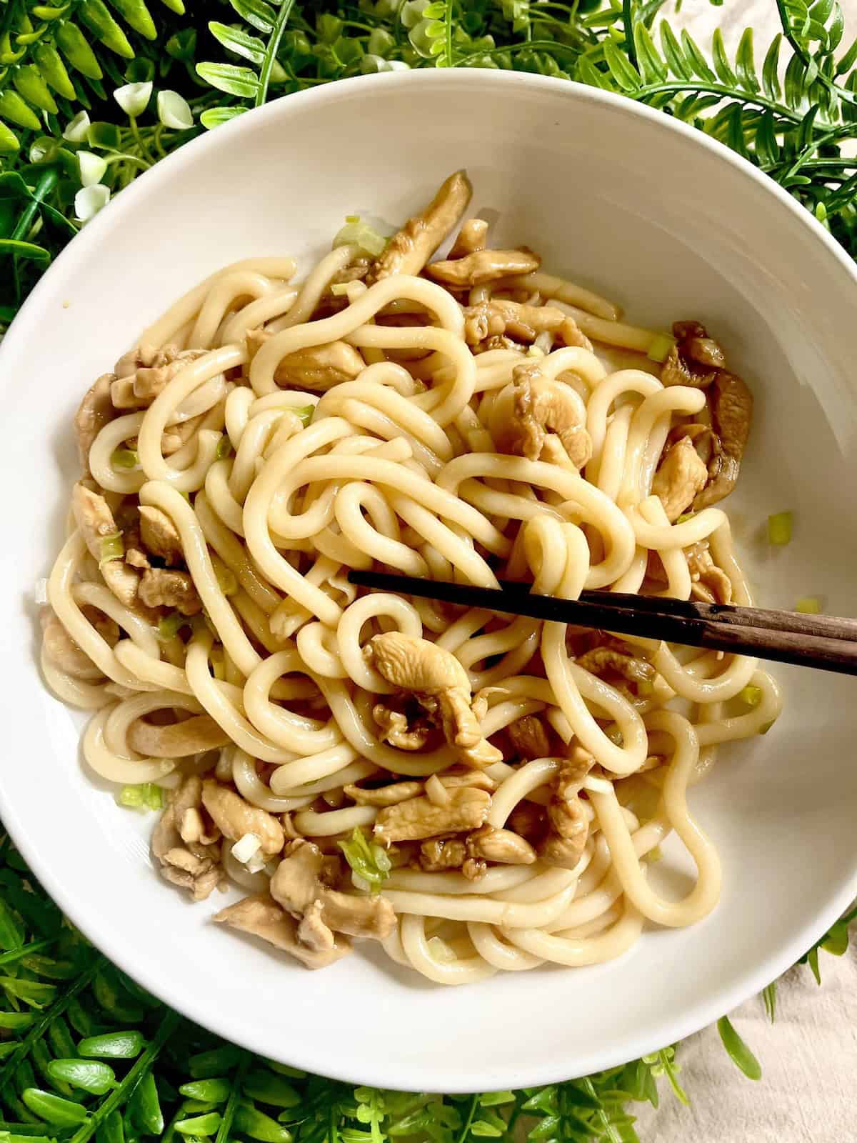 Chopsticks digging into a plate of stir-fried teriyaki noodles with chicken.