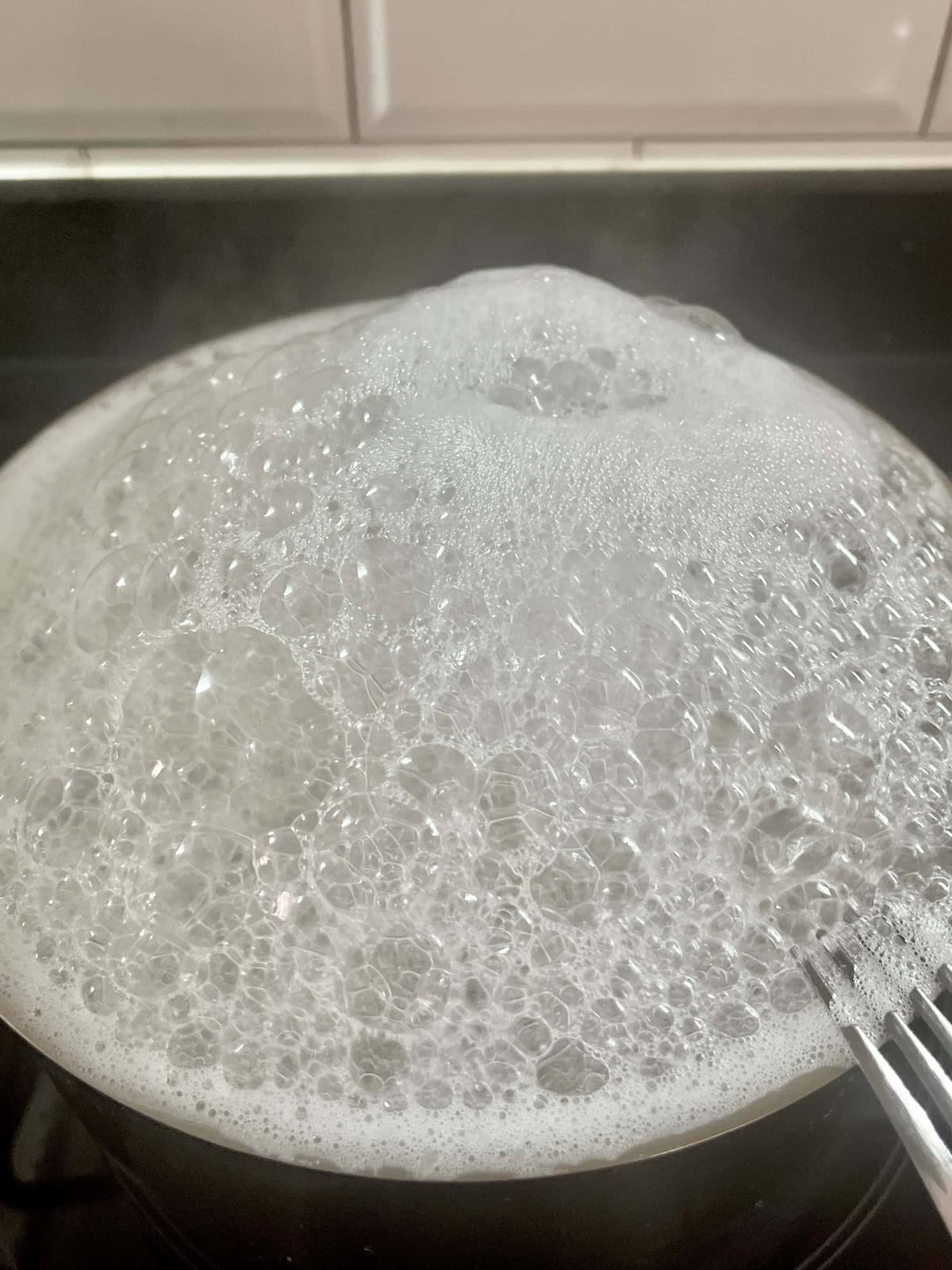 A pot of udon that is boiling over on the stove with lots of white foam.