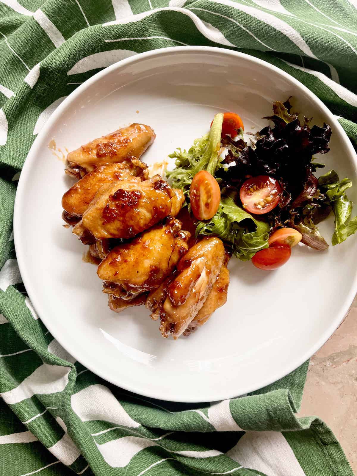 A pile of teriyaki chicken wings next to green salad on a white plate.