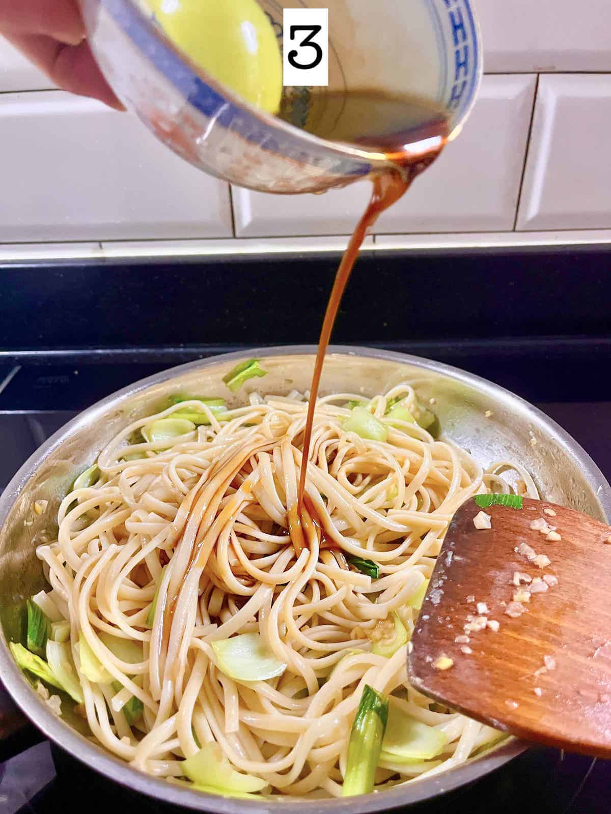 Pouring chow mein sauce over noodles in a silver pan.