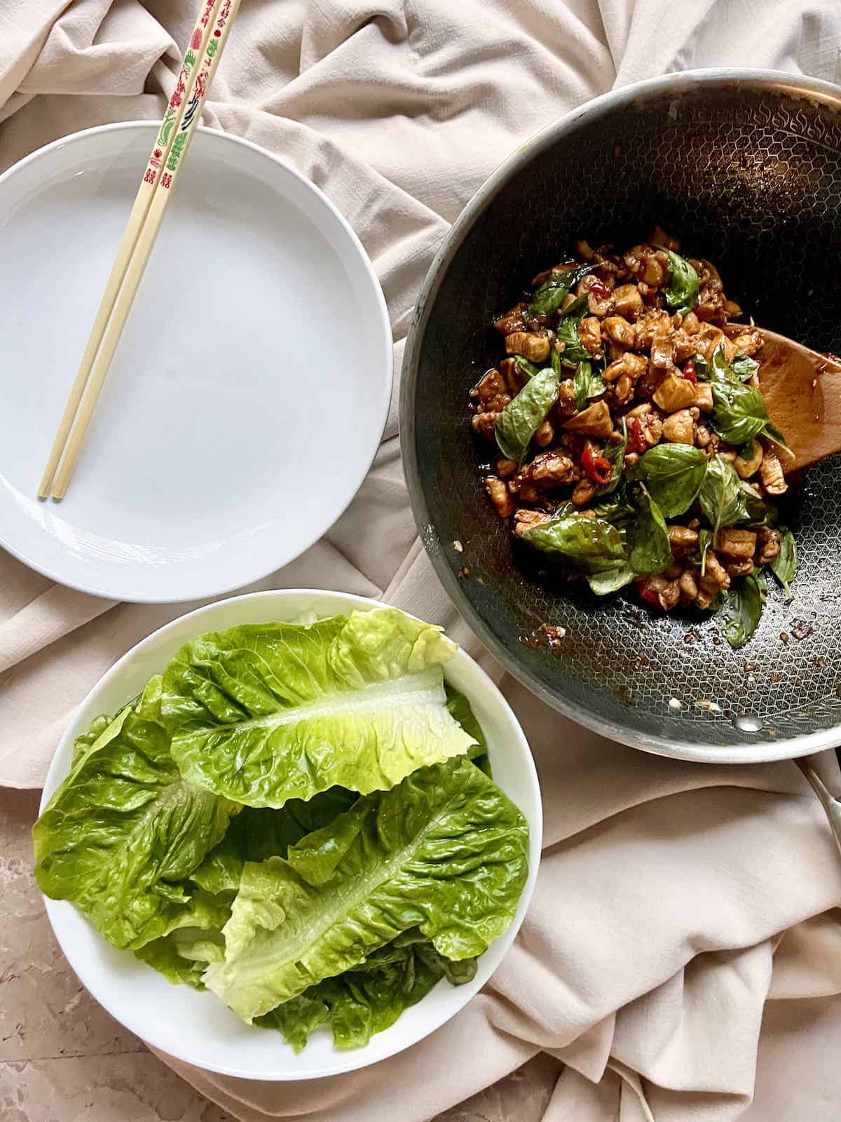 A pan full of Thai basil chicken stir fry and a bowl full of Romaine lettuce leaves.