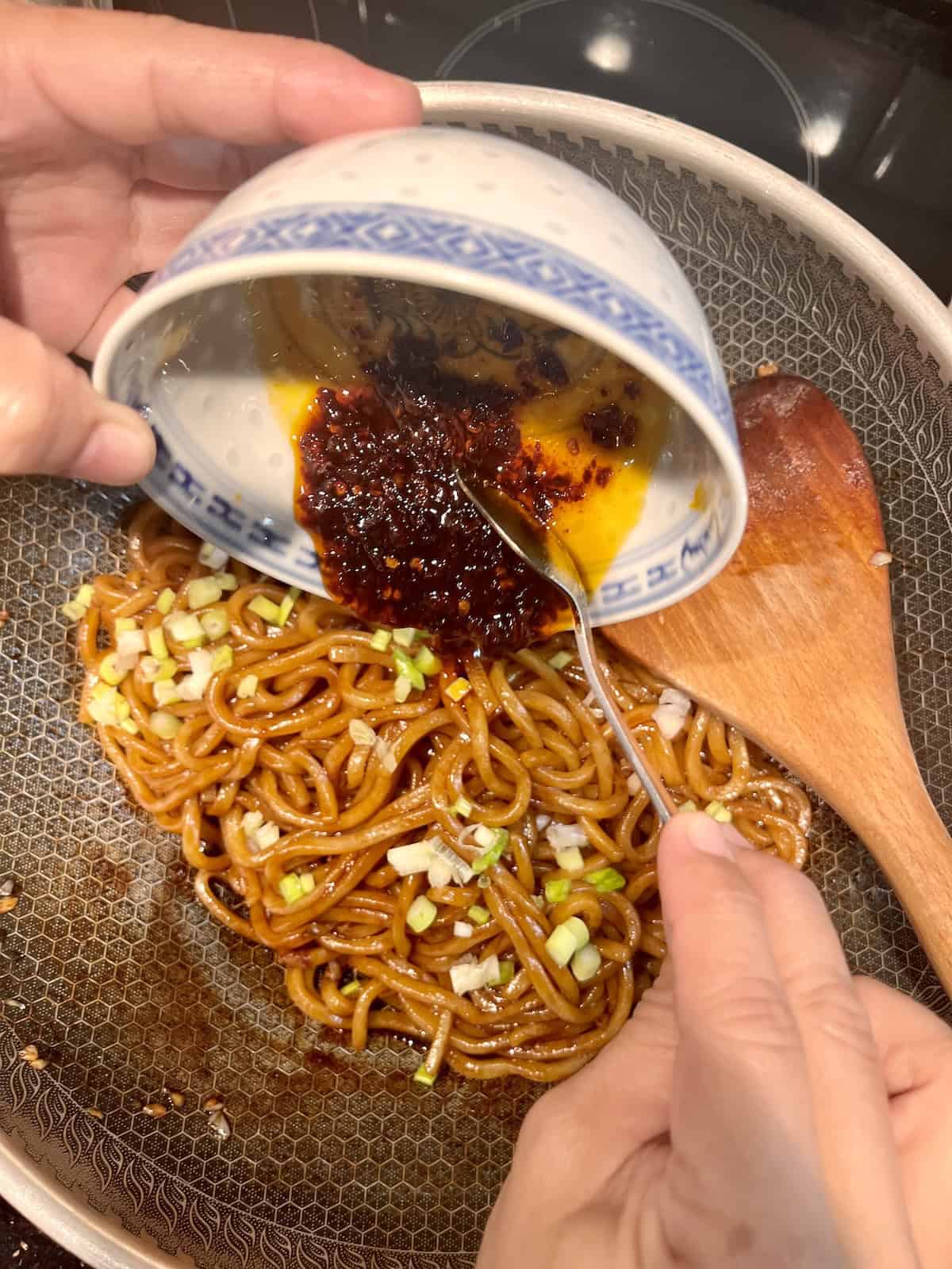 Pouring chili oil over udon noodles in a wok.